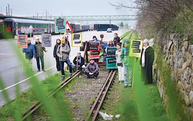 (Dieser ALT-Text wurde mithilfe von KI erstellt) Gruppe von Menschen steht und sitzt auf stillgelegten Bahngleisen und hält Schilder mit der Aufschrift „Westbahnpark jetzt!“. Im Hintergrund sind Züge zu sehen.