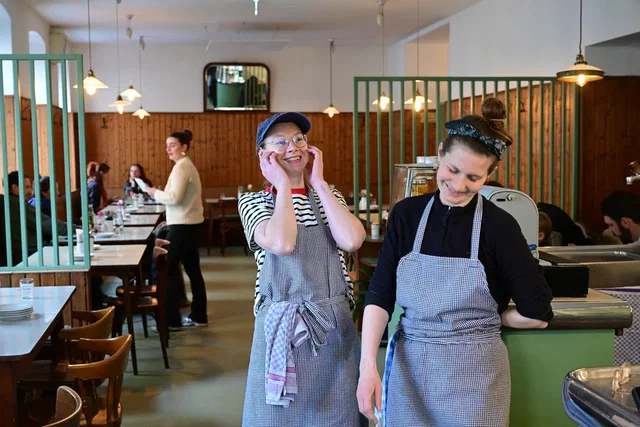 (Dieser ALT-Text wurde mithilfe von KI erstellt) Zwei Servicekräfte in Schürzen stehen lächelnd im Vordergrund eines gut besuchten Restaurants mit Holzwänden und gedeckten Tischen.