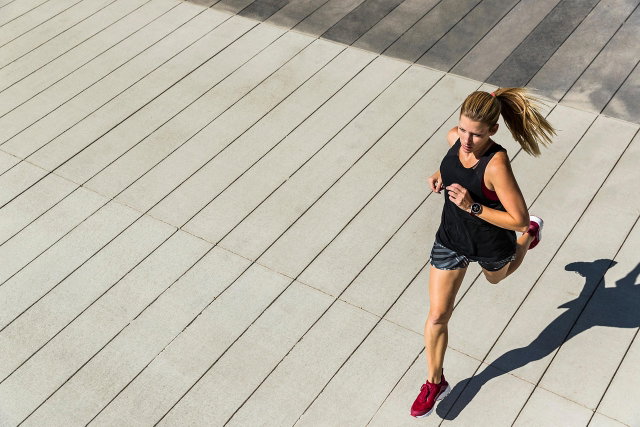 (Dieser ALT-Text wurde mithilfe von KI erstellt) Frau joggt auf hellem, gestreiftem Boden im Freien, trägt Sportkleidung und rote Laufschuhe, fotografiert aus der Vogelperspektive.