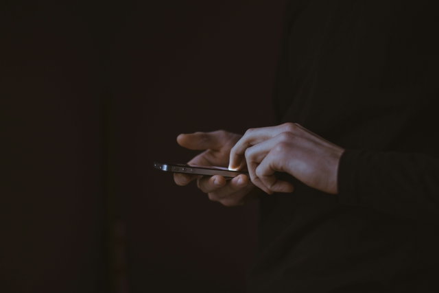 (Dieser ALT-Text wurde mithilfe von KI erstellt) Zwei Hände halten und bedienen ein Smartphone vor dunklem Hintergrund.