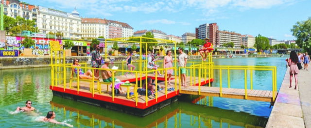 (Dieser ALT-Text wurde mithilfe von KI erstellt) Menschen entspannen und baden an einer gelben Badeplattform im Kanal einer Stadt mit umliegenden Gebäuden.