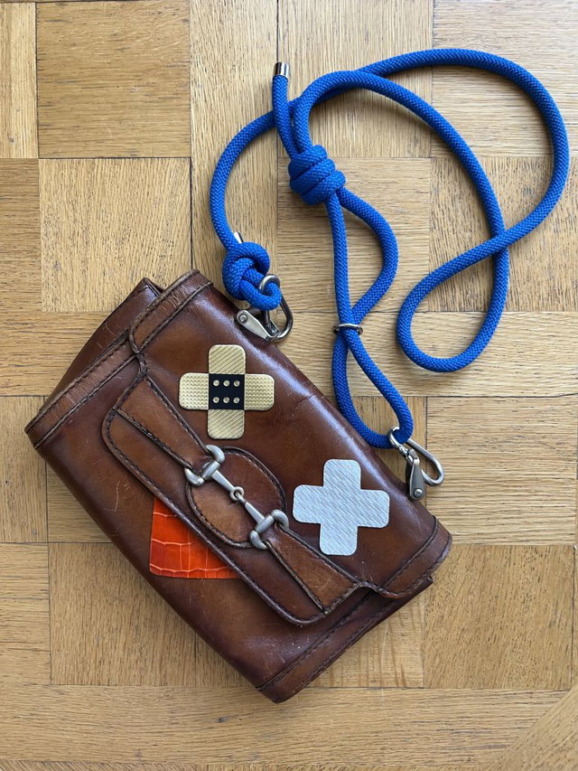 (Dieser ALT-Text wurde mithilfe von KI erstellt) Braune Ledertasche mit blauen Trageriemen, Pflaster- und Kreuzsymbol auf der Vorderseite, auf Holzoberfläche liegend.