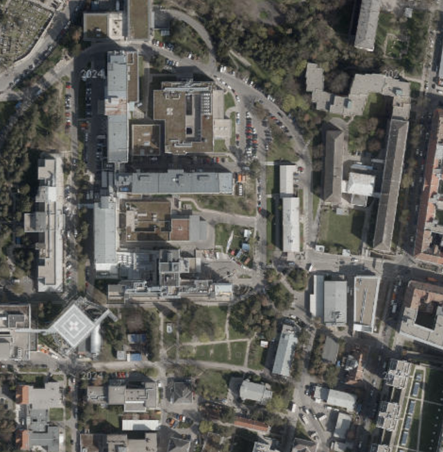 (Dieser ALT-Text wurde mithilfe von KI erstellt) Luftbildaufnahme eines städtischen Krankenhausgeländes mit mehreren Gebäuden, Straßen, Grünflächen und einem Hubschrauberlandeplatz.