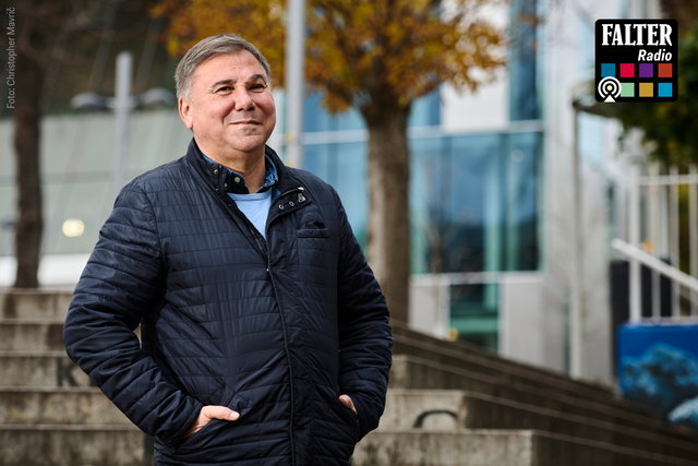 (Dieser ALT-Text wurde mithilfe von KI erstellt) Mann in dunkler Jacke steht draußen vor Treppen und modernen Gebäuden; oben rechts das Falter Radio Logo.