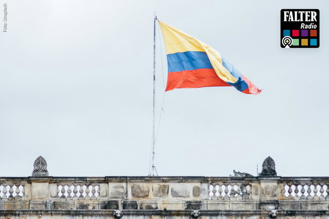 (Dieser ALT-Text wurde mithilfe von KI erstellt) Die kolumbianische Flagge weht auf einem Gebäude mit steinerner Balustrade, oben rechts das Falter Radio Logo.