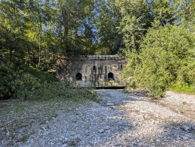 (Dieser ALT-Text wurde mithilfe von KI erstellt) Trockenes Flussbett mit wenigen Wasserresten vor einem alten, überwucherten Betonbauwerk im Wald.