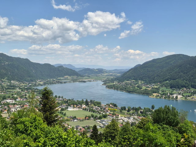 (Dieser ALT-Text wurde mithilfe von KI erstellt) Blick auf den Ossiacher See mit umliegenden Ortschaften, bewaldeten Bergen und teils bewölktem Himmel.